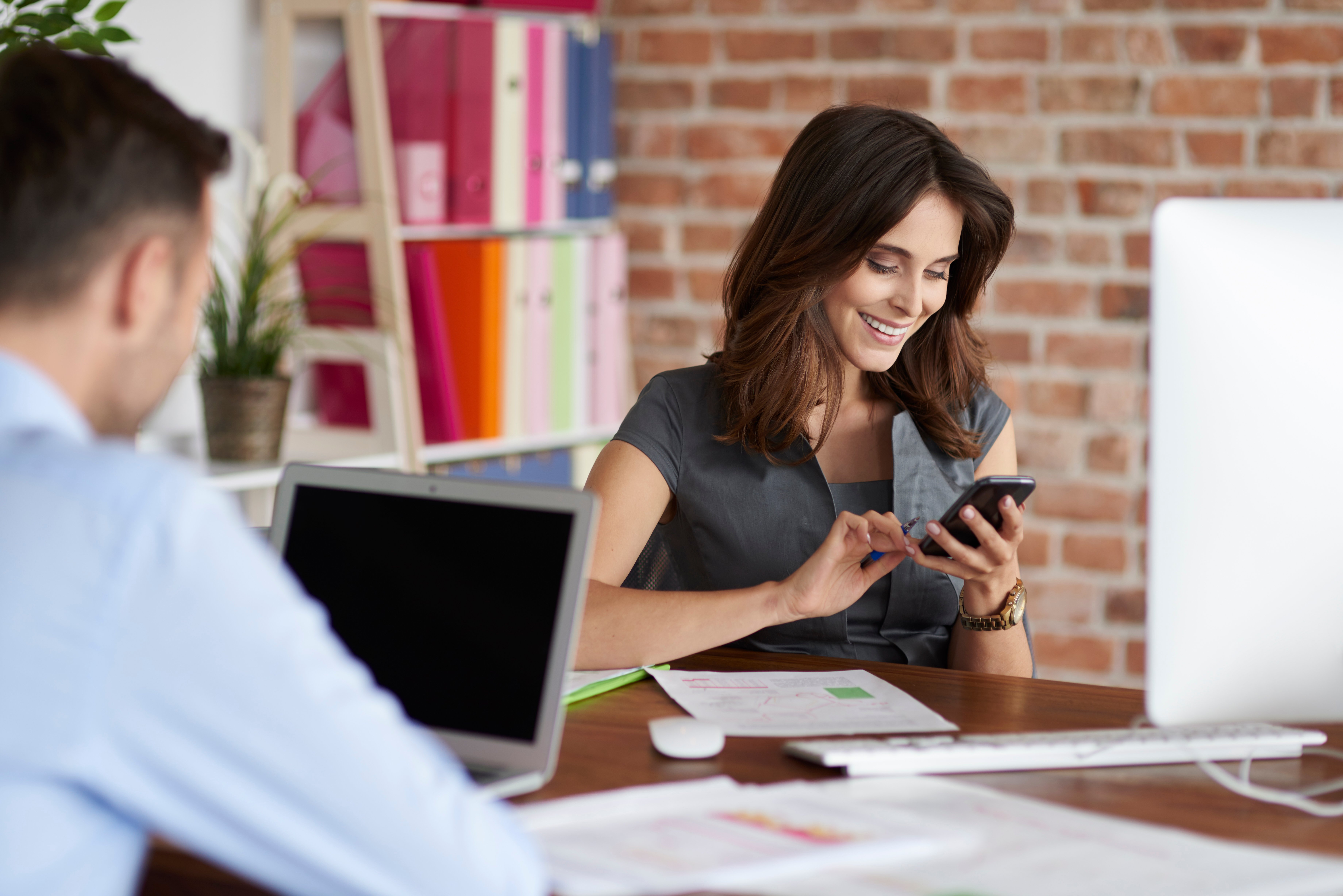 A smiling woman checks her phone at a desk while a colleague works nearby in an office.